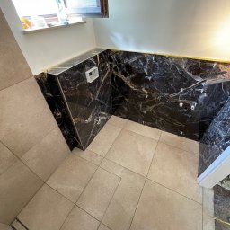 Mateusz Leśnikowski - Wykończona łazienka z czarnymi płytkami imitującymi marmur na ścianach i beżowymi płytkami na podłodze. Widoczne elementy instalacji wodnej i okno w tle.