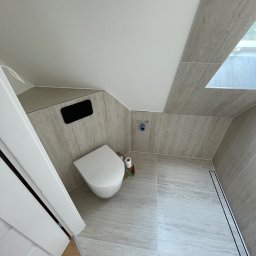 Mateusz Leśnikowski - Nowoczesna łazienka na poddaszu z beżowymi płytkami imitującymi kamień, wiszącą toaletą i odpływem liniowym. Ujęcie z góry podkreśla skosy i okno dachowe.