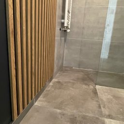 Mateusz Leśnikowski - Nowoczesna łazienka z drewnianą ścianką działową, szarymi płytkami i grzejnikiem drabinkowym. Minimalistyczny design, widoczny fragment kabiny prysznicowej.