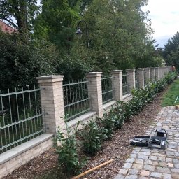 Mateusz Leśnikowski - Murowane ogrodzenie z metalowymi przęsłami, obsadzone roślinami, widoczne narzędzia pracy na brukowanej ścieżce. Praca w toku, świeżo posadzone krzewy.