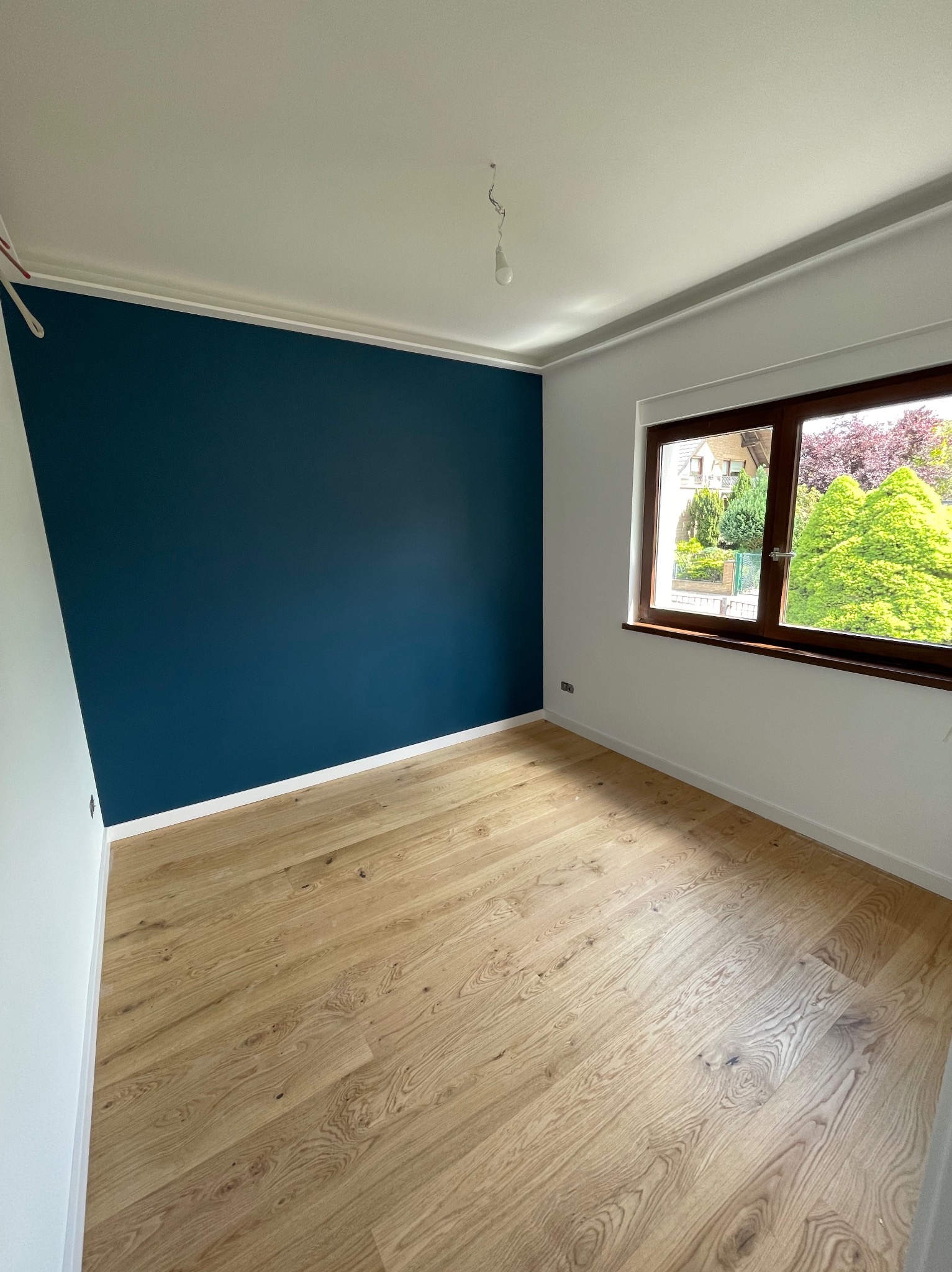 Pomalowany na granatowo fragment ściany w pokoju z jasną podłogą i białymi ścianami, widok na zieleń za oknem. Listwy przypodłogowe i sufitowe w kolorze białym.