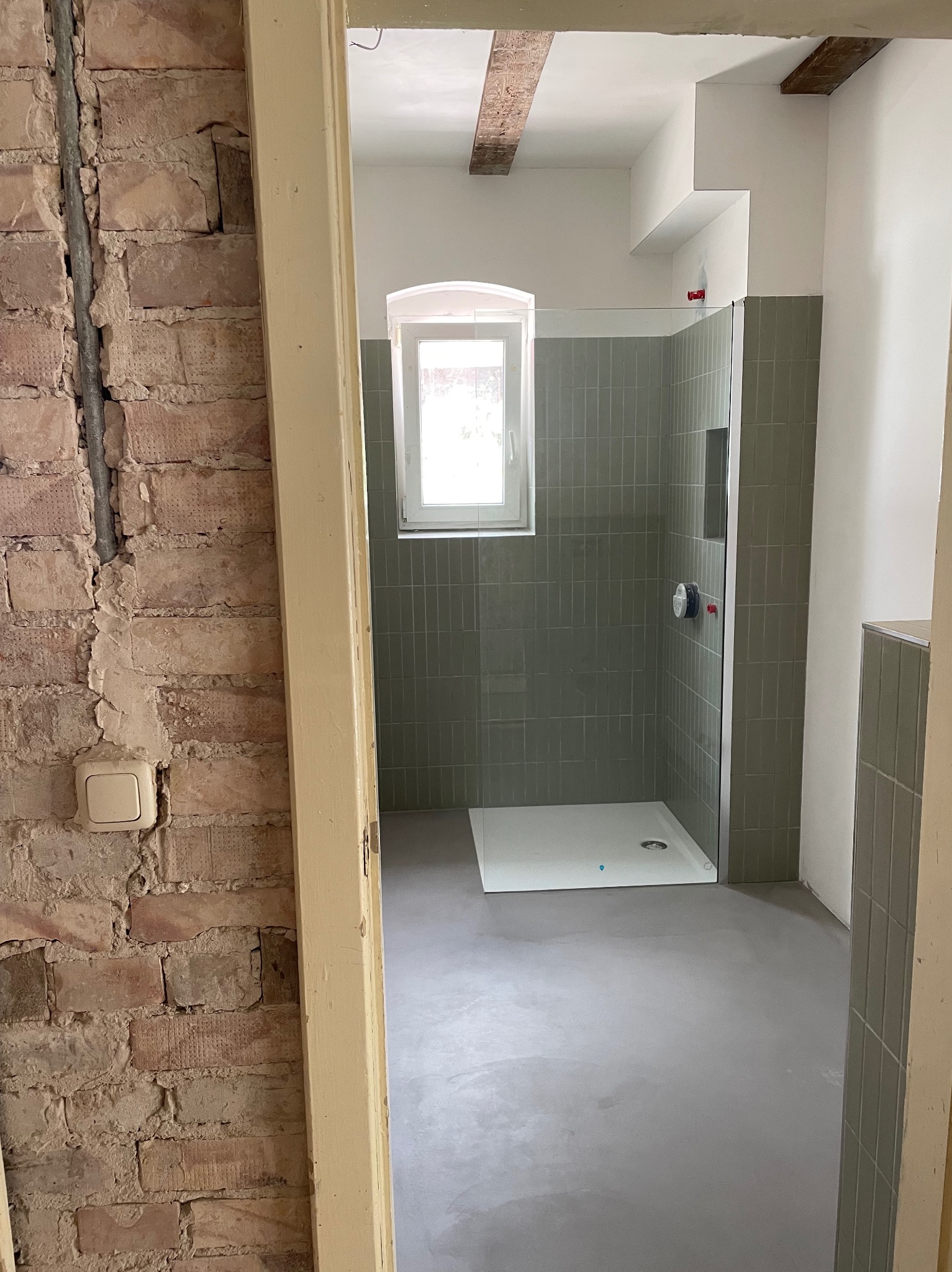 Łazienka w trakcie remontu: surowa cegła kontrastuje z nową kabiną prysznicową i szarym betonem na podłodze. Widoczne elementy instalacji i wykończenia.