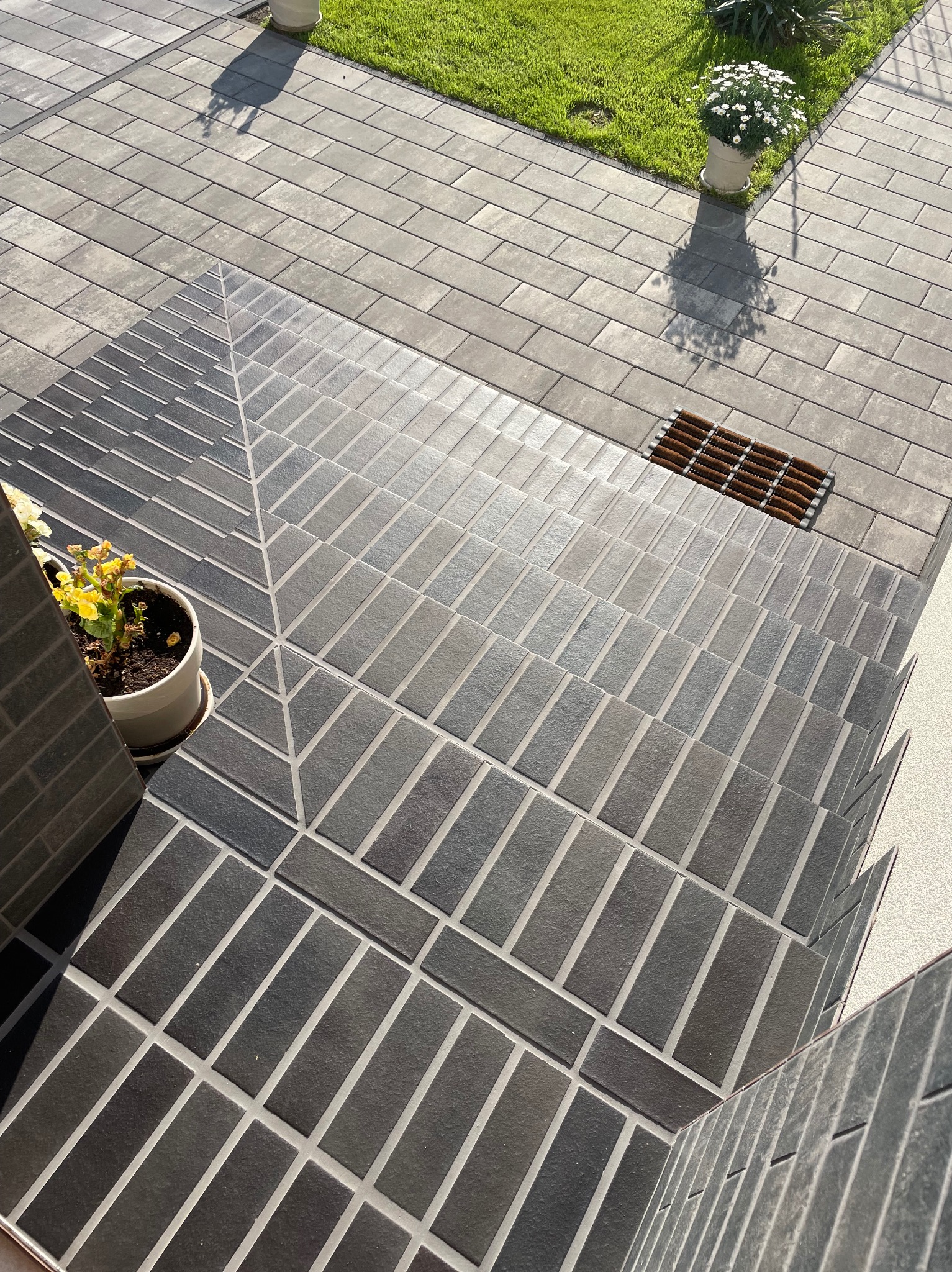 Schody wejściowe wyłożone szarą, prostokątną kostką brukową z jasnymi fugami, prowadzące do trawnika z kwiatami w donicach. Widok z góry, dywanik przed drzwiami.