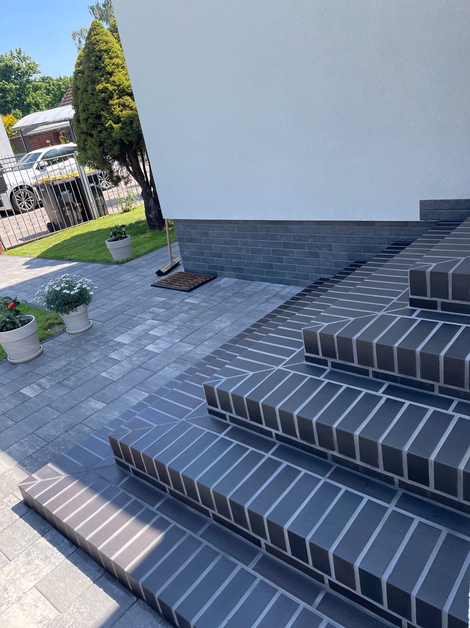 Schody wejściowe wyłożone ciemną, ceglastą kostką brukową, prowadzące do białego budynku. Widoczne donice z kwiatami i fragment posesji z samochodem.