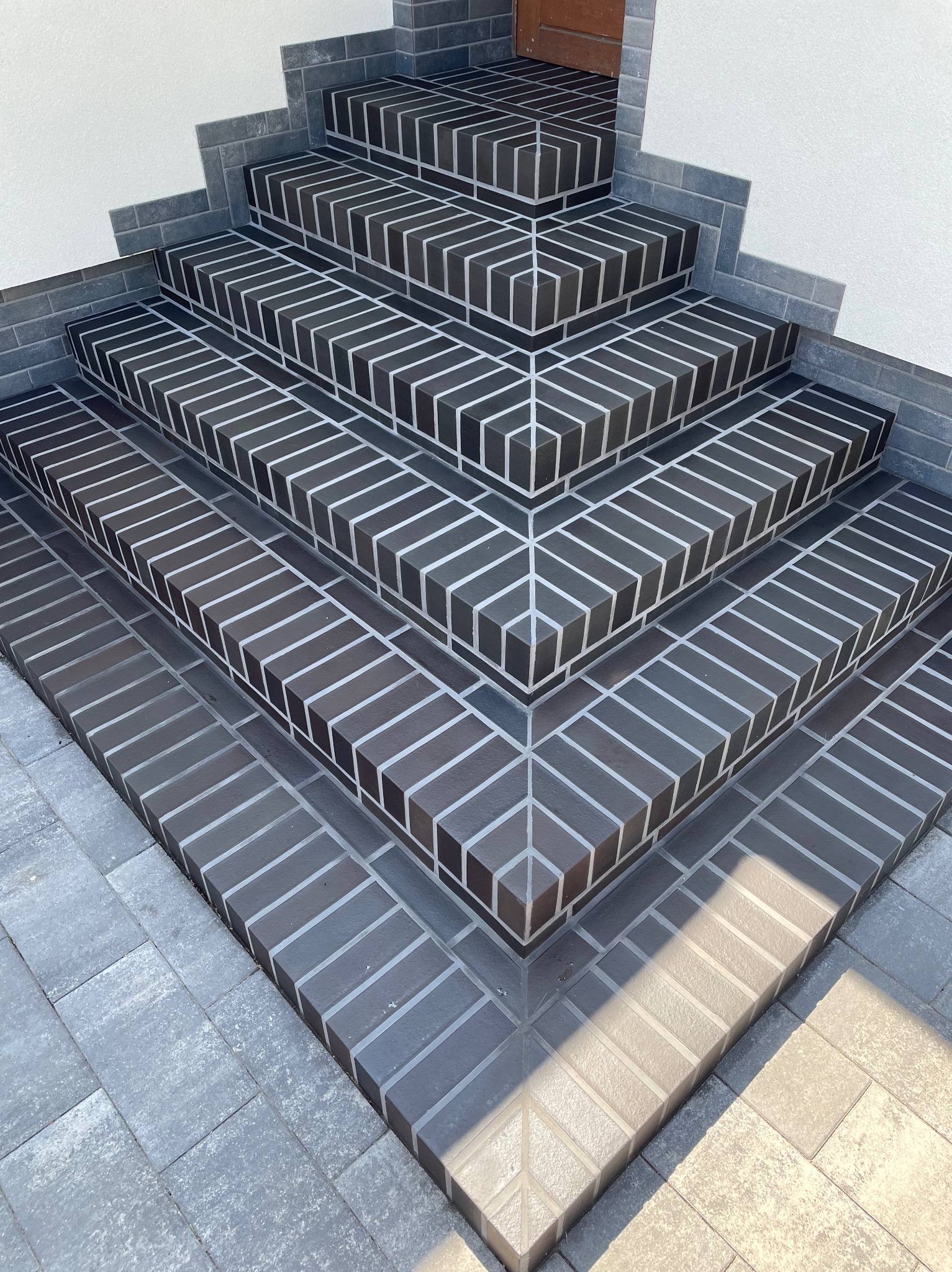 Schody wejściowe wyłożone ciemną cegłą klinkierową z jasną fugą, tworzące geometryczny wzór. Widoczne detale wykończenia i połączenie z szarą kostką brukową.