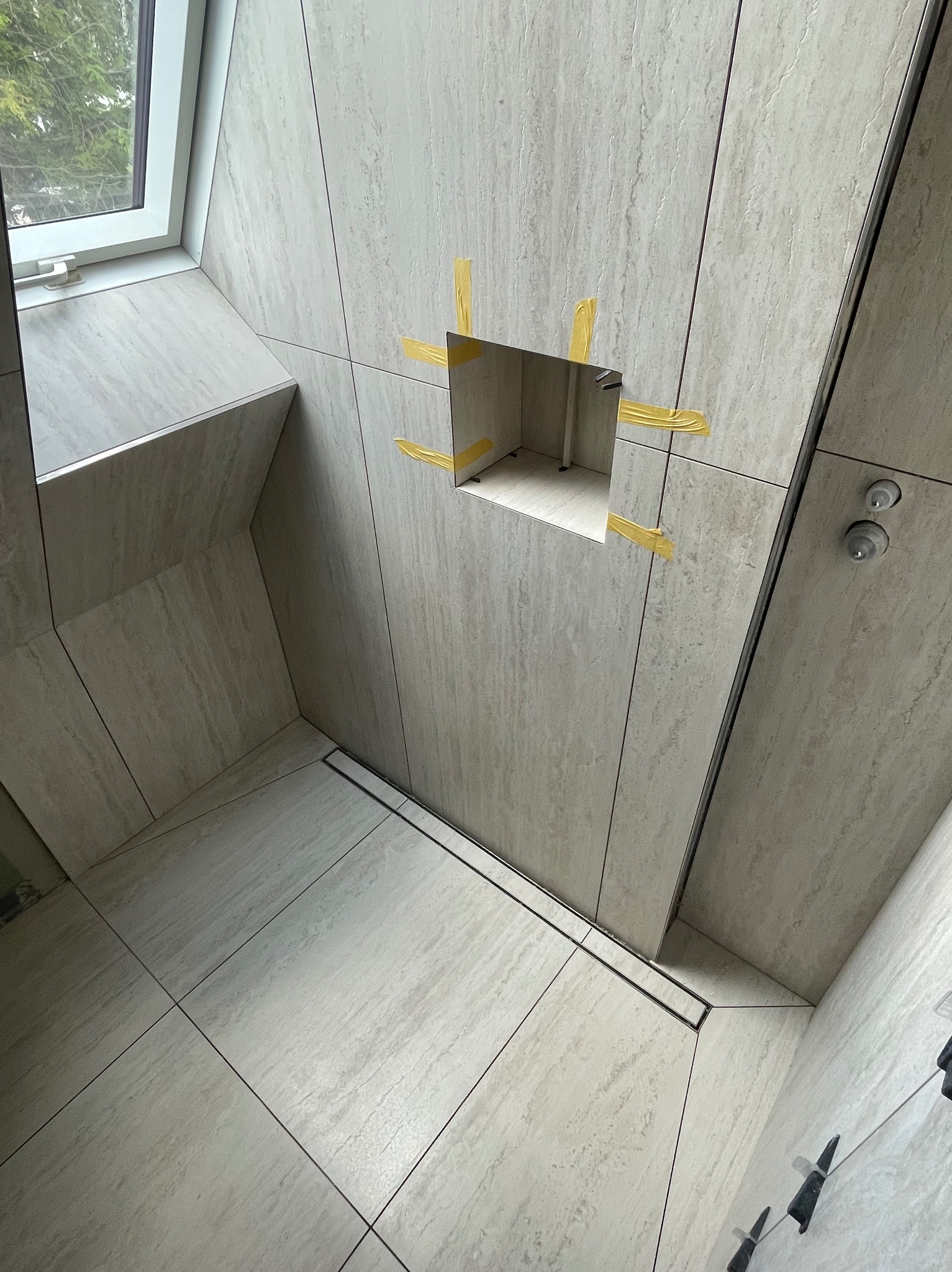 Wykończona łazienka z beżowymi płytkami imitującymi kamień. Widoczny odpływ liniowy, okno dachowe i otwór na baterię zabezpieczony taśmą. Minimalistyczny design.