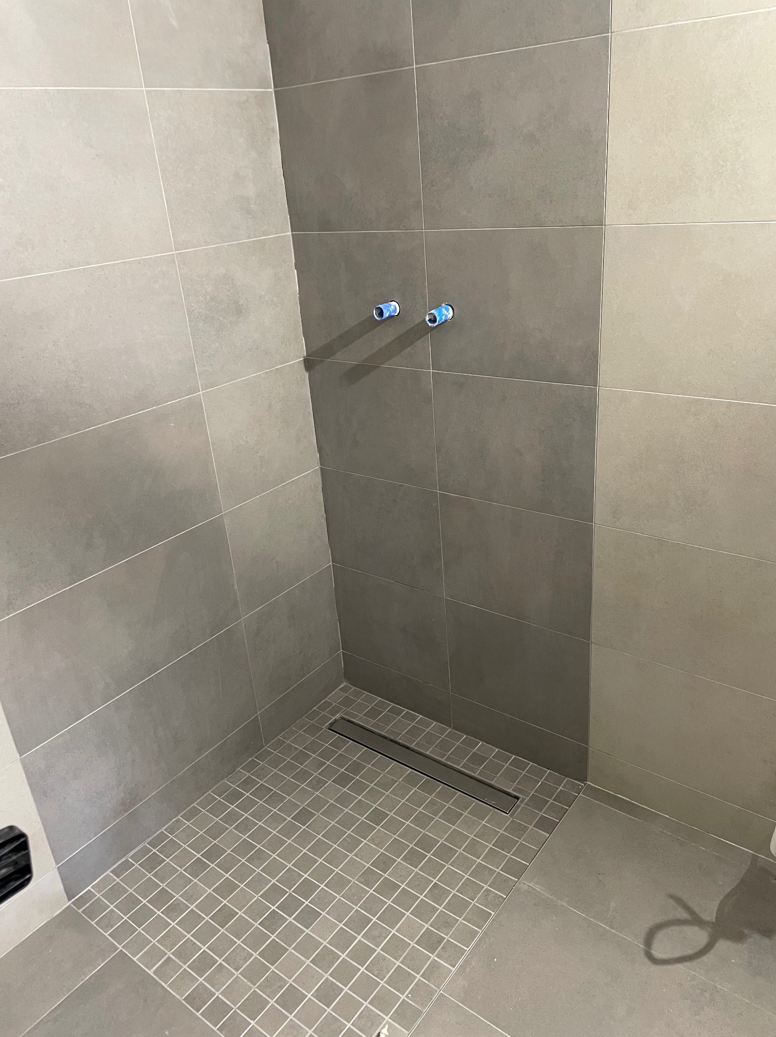 Szara, wykafelkowana kabina prysznicowa z odpływem liniowym i wystającymi rurami instalacji wodnej. Minimalistyczny design, neutralna kolorystyka.