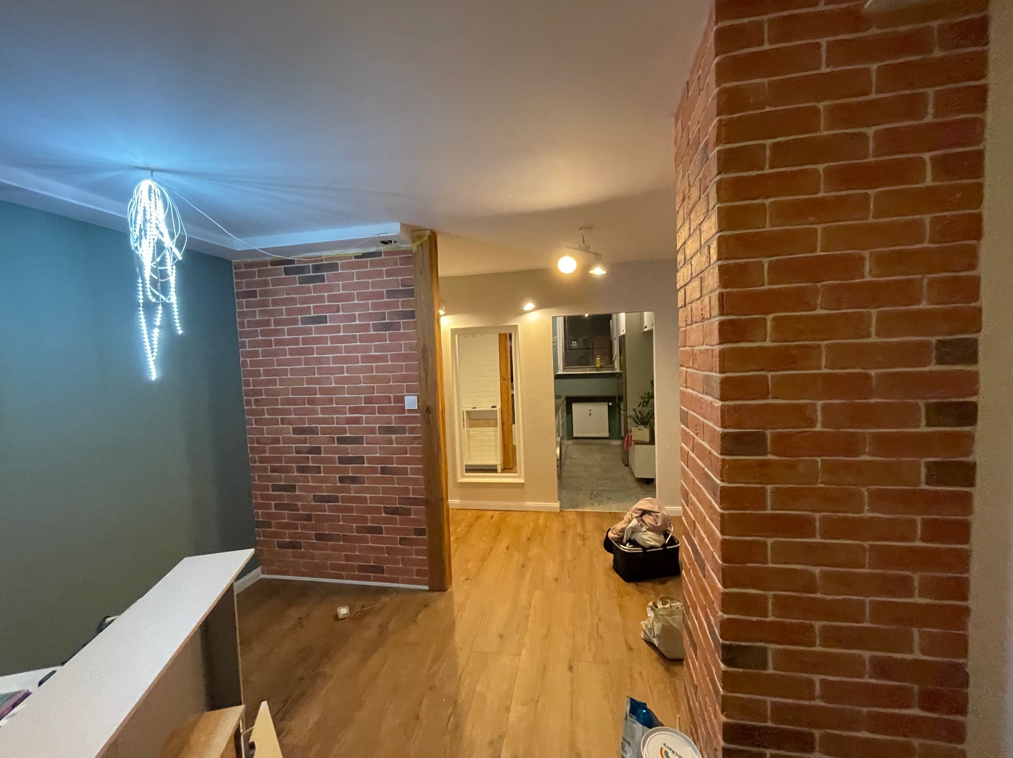 Ceglana ściana działowa w odcieniach czerwieni, drewniana podłoga, widok na dalsze pomieszczenia. Dekoracyjne oświetlenie sufitowe. Częściowo widoczne przedmioty w pomieszczeniu.