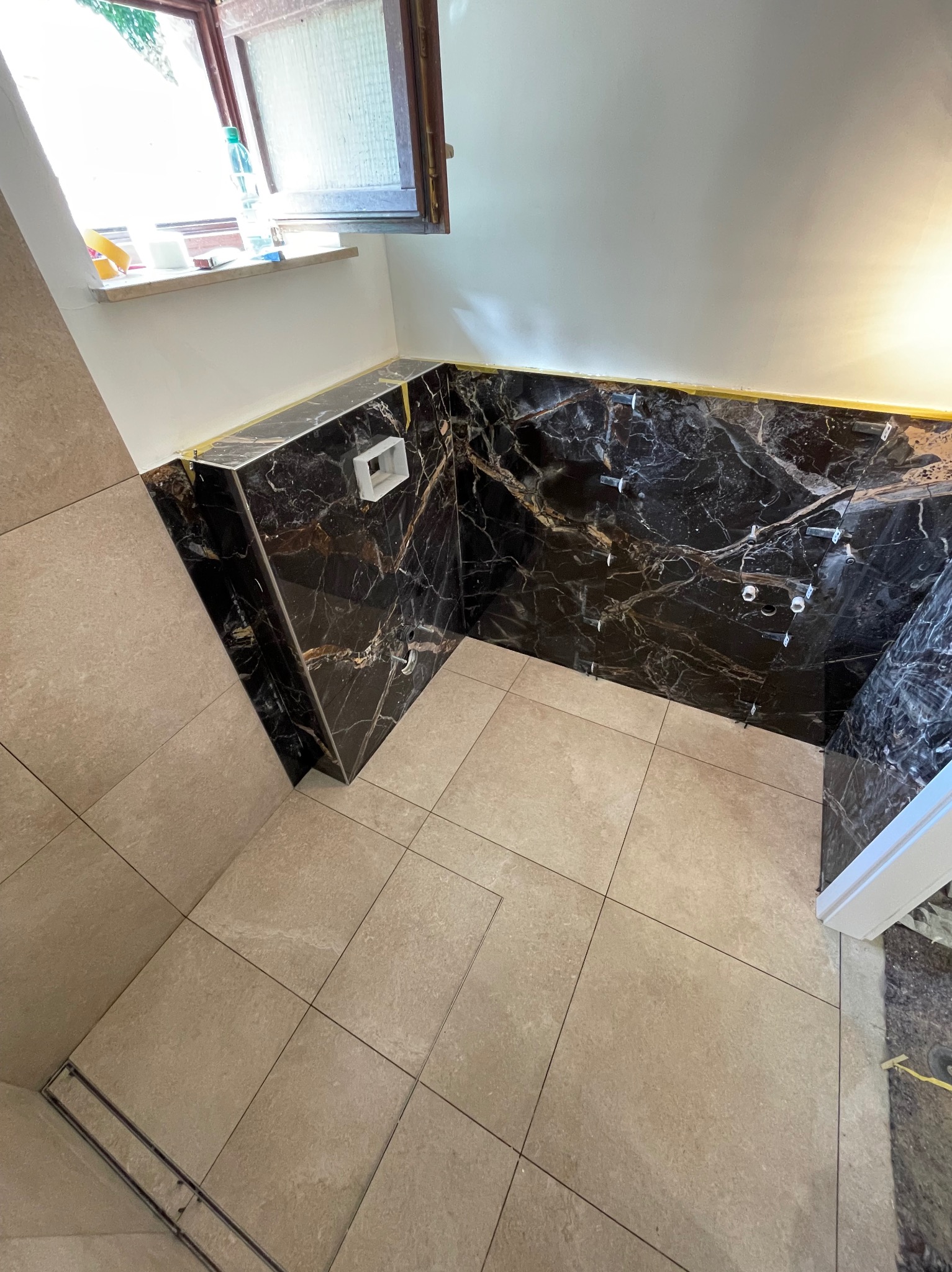 Wykończona łazienka z czarnymi płytkami imitującymi marmur na ścianach i beżowymi płytkami na podłodze. Widoczne elementy instalacji wodnej i okno w tle.