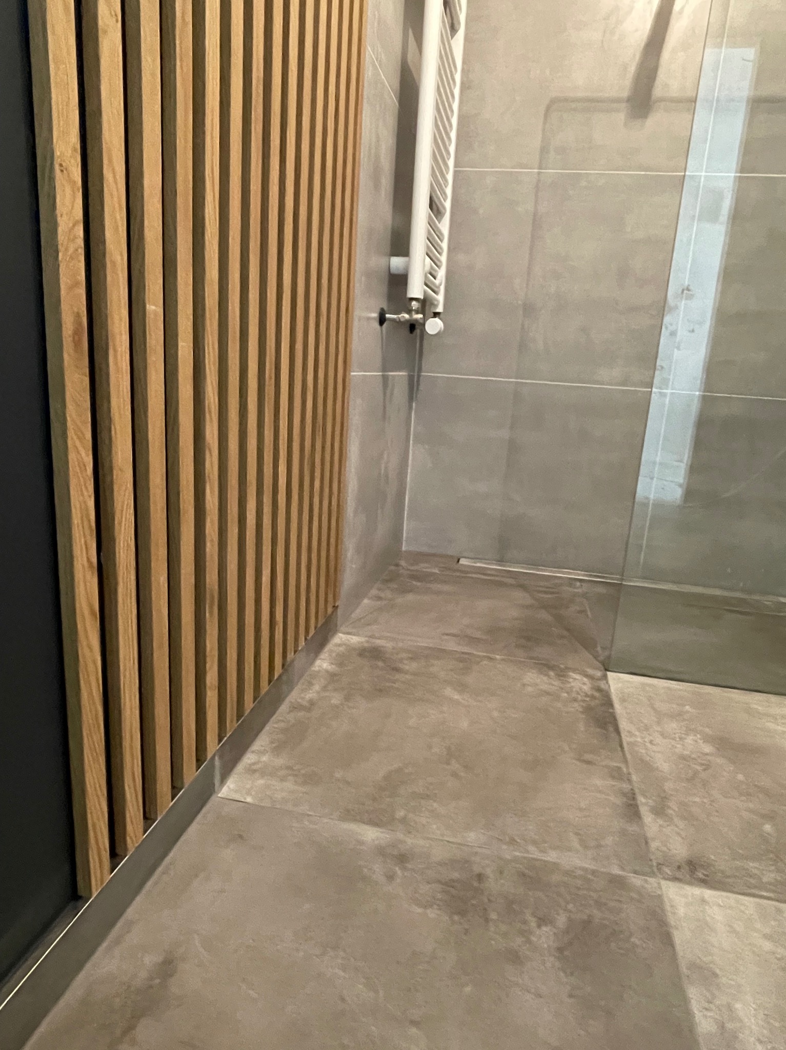 Nowoczesna łazienka z drewnianą ścianką działową, szarymi płytkami i grzejnikiem drabinkowym. Minimalistyczny design, widoczny fragment kabiny prysznicowej.