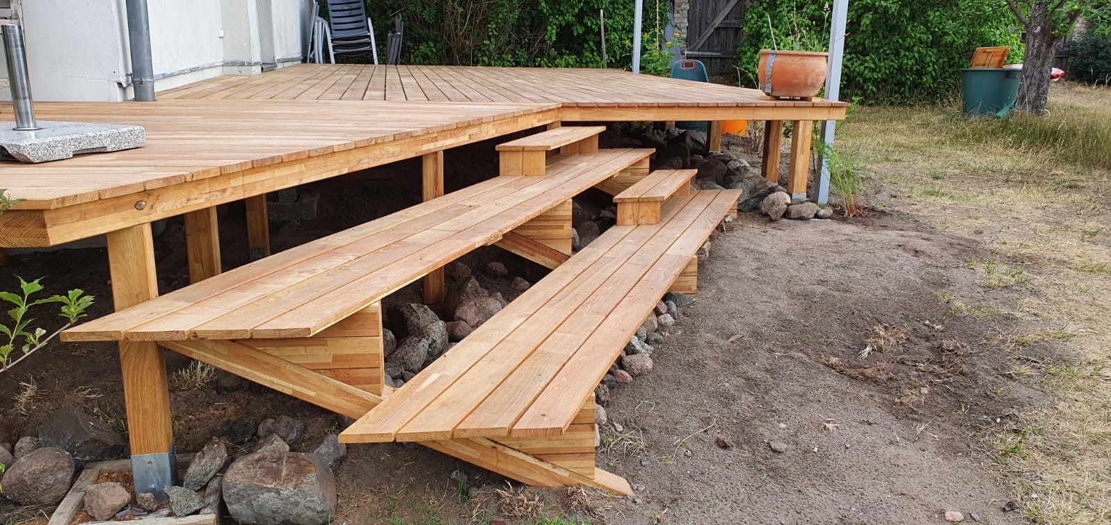 Drewniany taras z schodami na zewnątrz, wykonany z jasnego drewna, z widocznymi kamieniami pod konstrukcją. W tle zieleń i elementy architektury.