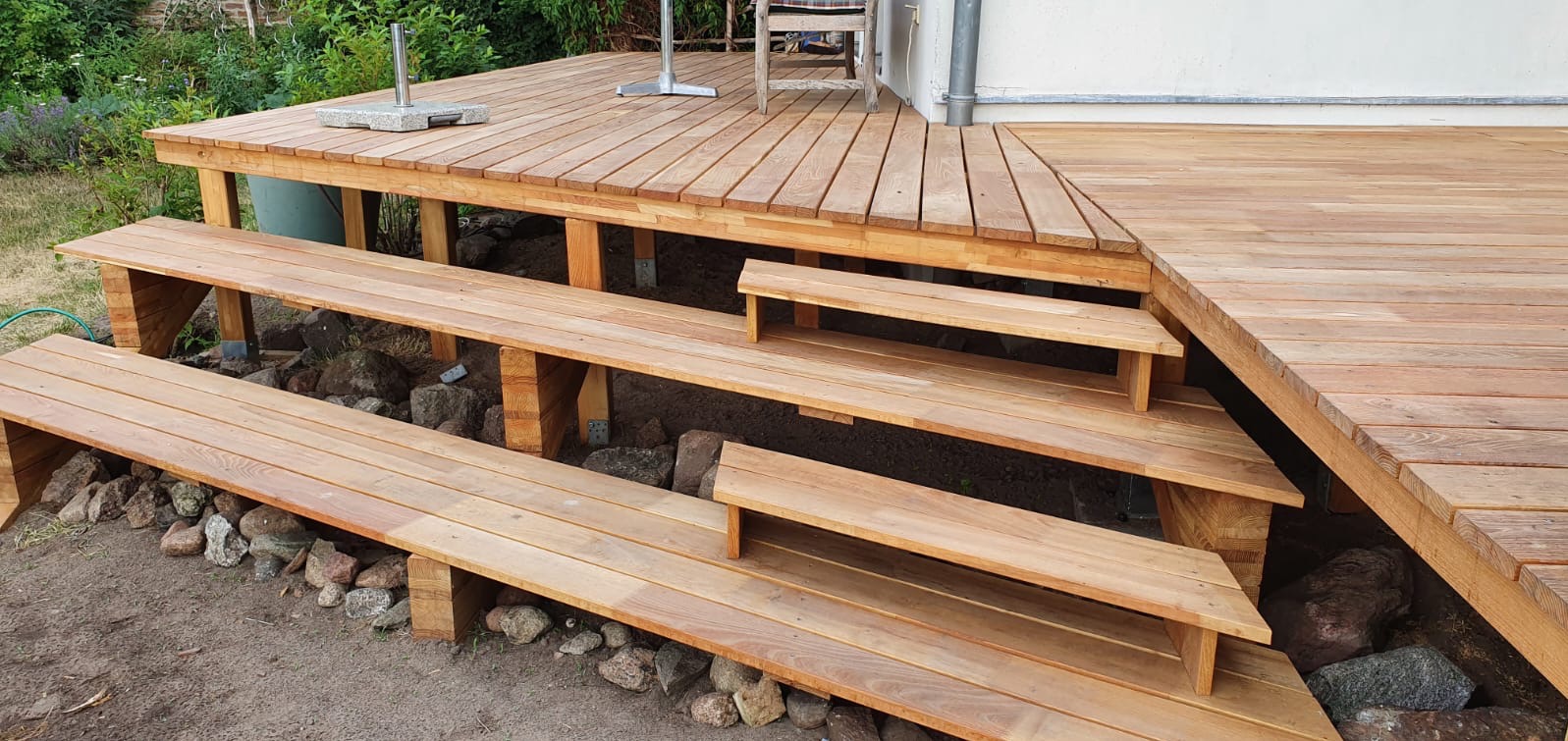 Drewniany taras z ławkami na nierównym terenie, widoczne kamienie i zieleń w tle. Jasne drewno, schody prowadzące na taras, parasol ogrodowy.