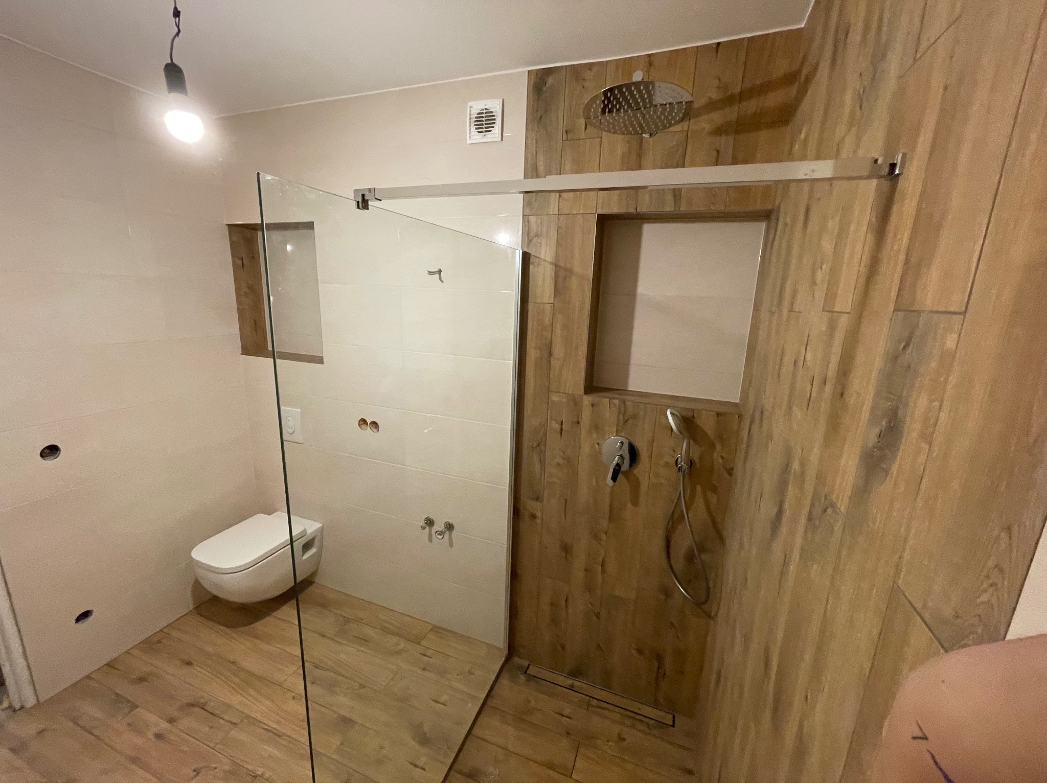 Nowoczesna łazienka z kabiną prysznicową typu walk-in, wykończona płytkami imitującymi drewno i białymi płytkami. Widoczna toaleta podwieszana i deszczownica.