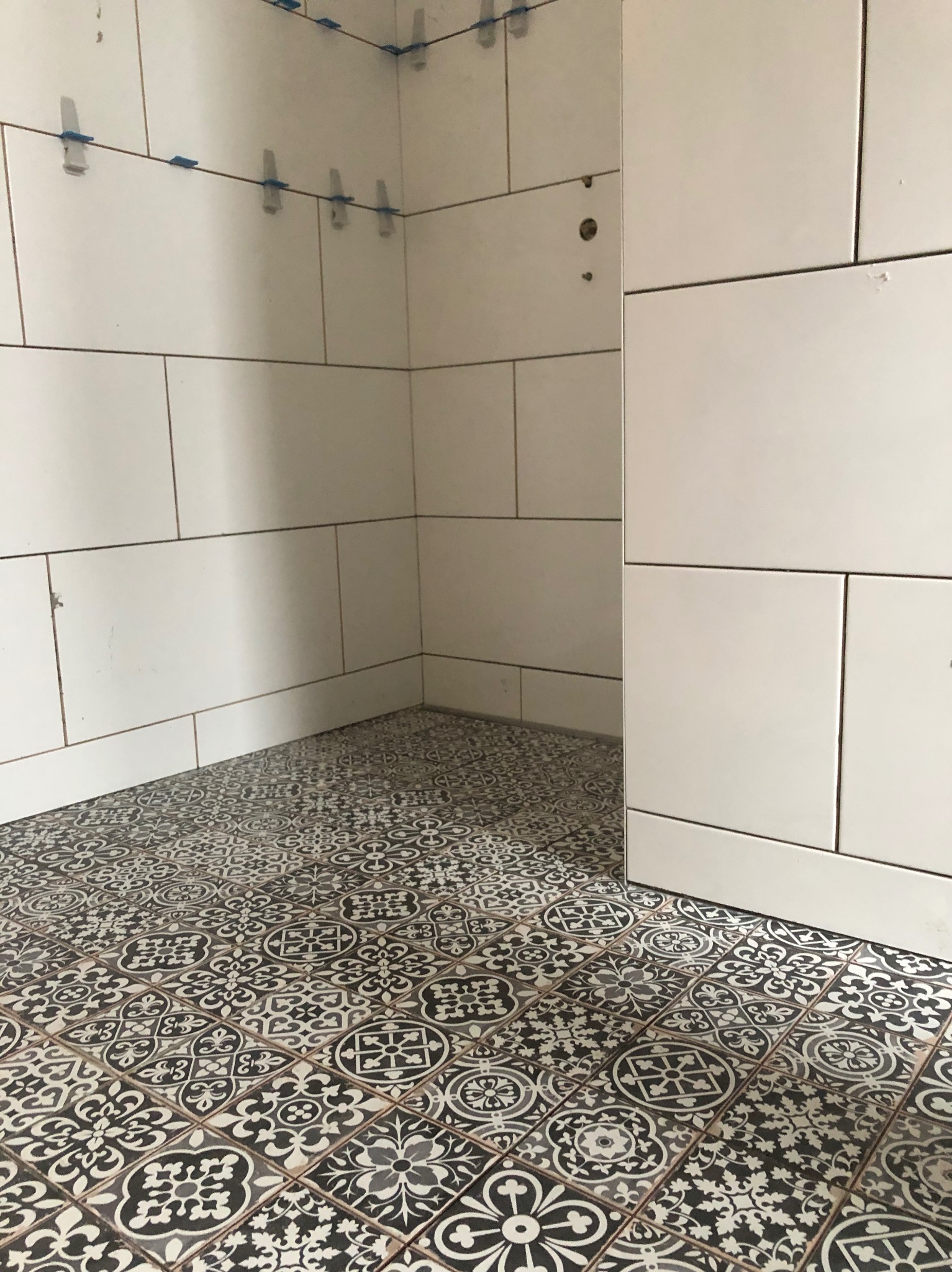 Białe płytki ścienne z niebieskimi krzyżykami poziomującymi i wzorzysta, czarno-biała podłoga z płytek w łazience w trakcie wykańczania.