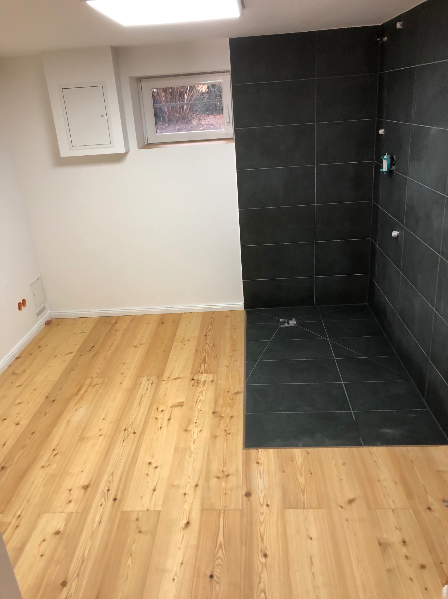Nowoczesna łazienka w trakcie wykończenia: drewniana podłoga kontrastuje z czarnymi płytkami w kabinie prysznicowej. Widoczne okno i dostęp do instalacji.