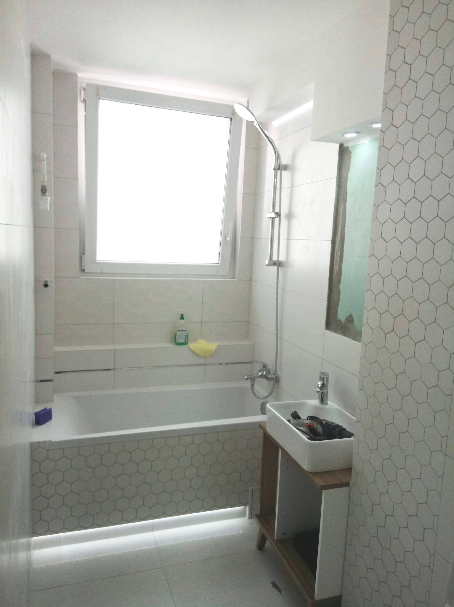 Jasna łazienka po remoncie: wanna obudowana heksagonalnymi płytkami, umywalka nablatowa na drewnianym stelażu, białe płytki na ścianach i podłodze, okno.