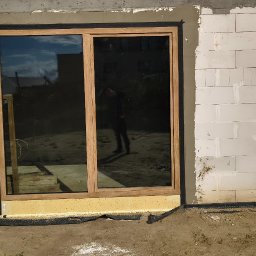 Przedstawiciel handlowy firmy Drutex ,Eko okna,i gdanskiej fabryki okien - Montaż okna tarasowego z drewnianą ramą w surowym murze z betonu komórkowego. Widoczne detale montażu i odbicie osoby w szybie.