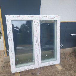 Przedstawiciel handlowy firmy Drutex ,Eko okna,i gdanskiej fabryki okien - Dwu-skrzydłowe okno PCV marki Aluplast, białe, leżące na drewnianych deskach na tle elewacji w trakcie montażu. Widoczne detale profili i odblask osoby robiącej zdjęcie.