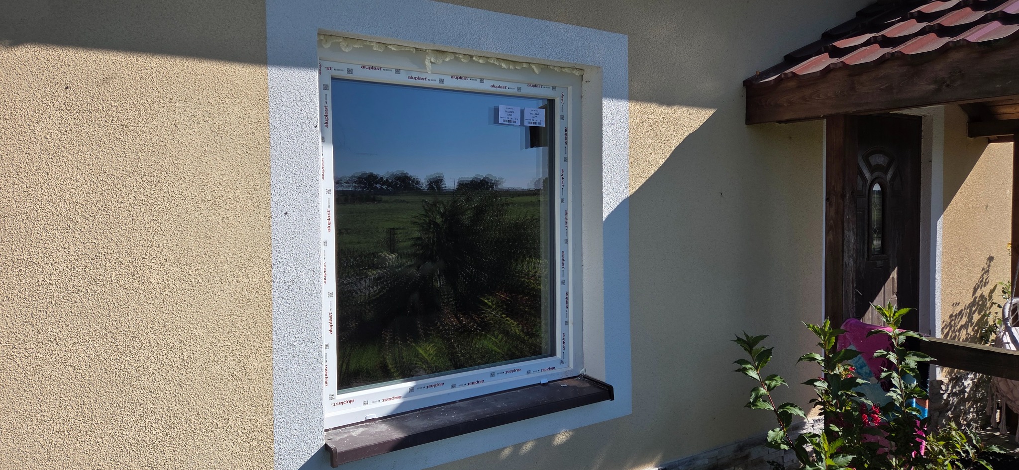 Białe okno PCV z widokiem na zewnątrz, osadzone w ścianie budynku z ociepleniem. Widoczne pianka montażowa i parapet zewnętrzny. Na szybie naklejki.