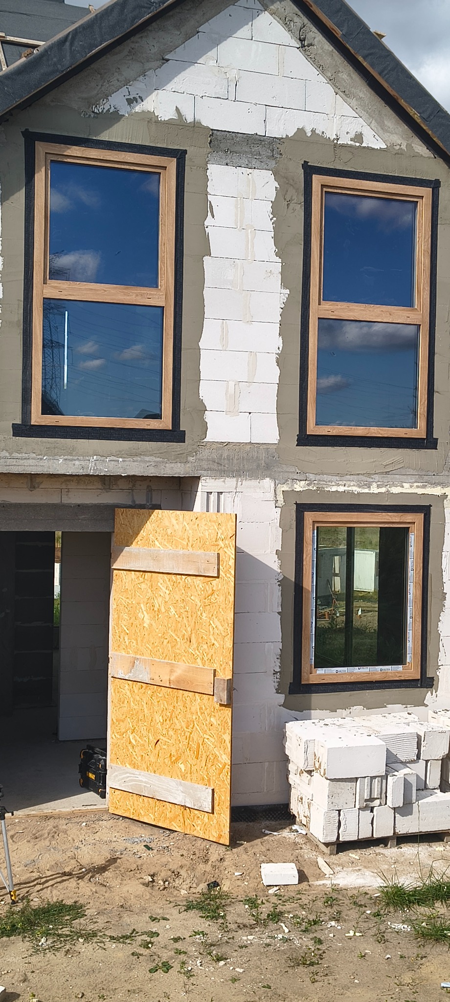 Montaż okien z drewnianą ramą w surowym budynku z białego betonu komórkowego. Na dole prowizoryczne drzwi z płyty OSB.