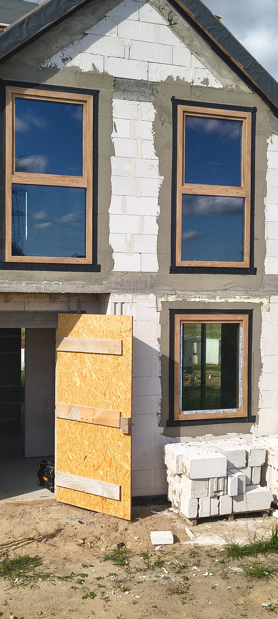 Montaż okien z drewnianą ramą w surowym budynku z białych bloczków. Widoczna tymczasowa osłona wejścia i materiały budowlane.