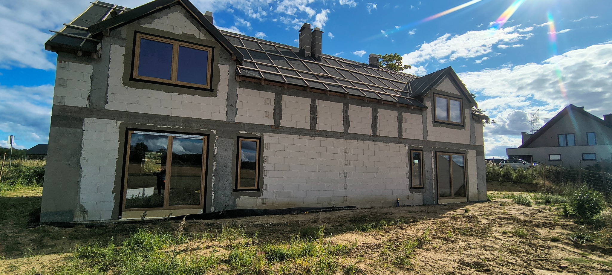 Surowy stan domu jednorodzinnego z zamontowanymi oknami w drewnianych ramach, widok z zewnątrz, na dachu widoczna konstrukcja pod dachówkę.