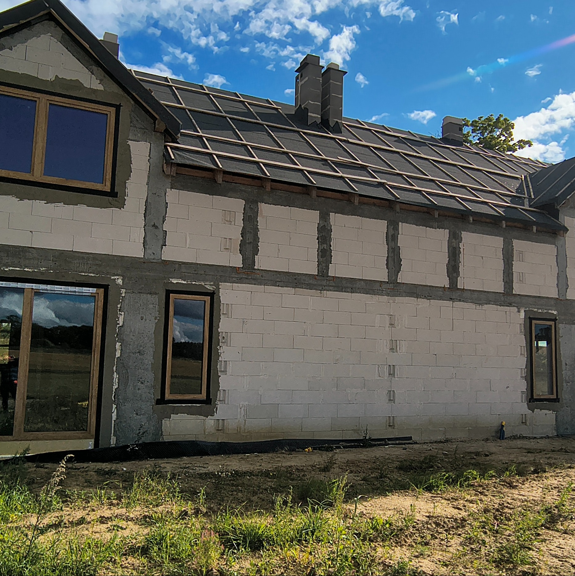 Budowa domu z oknami w drewnianej ramie i dachem z czarnej membrany, widok na elewację z bloczków, błękitne niebo.