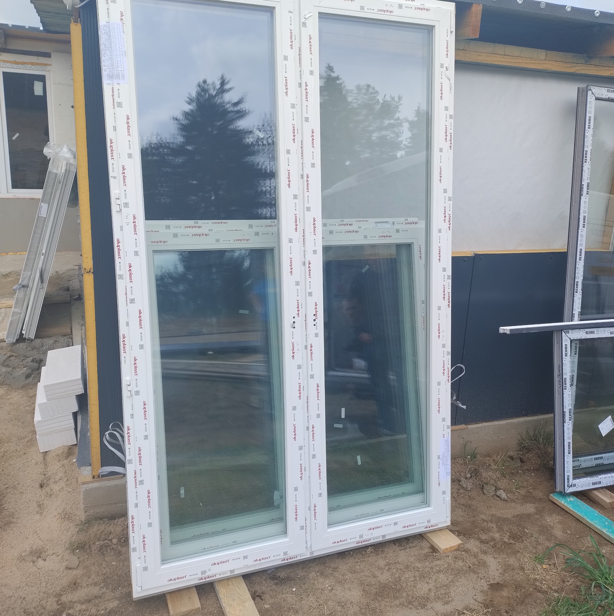 Dwuokienne, białe okno PCV Aluplast, jeszcze nie zamontowane, oparte o ścianę budynku w trakcie budowy. Widoczne bloczki i drabina.