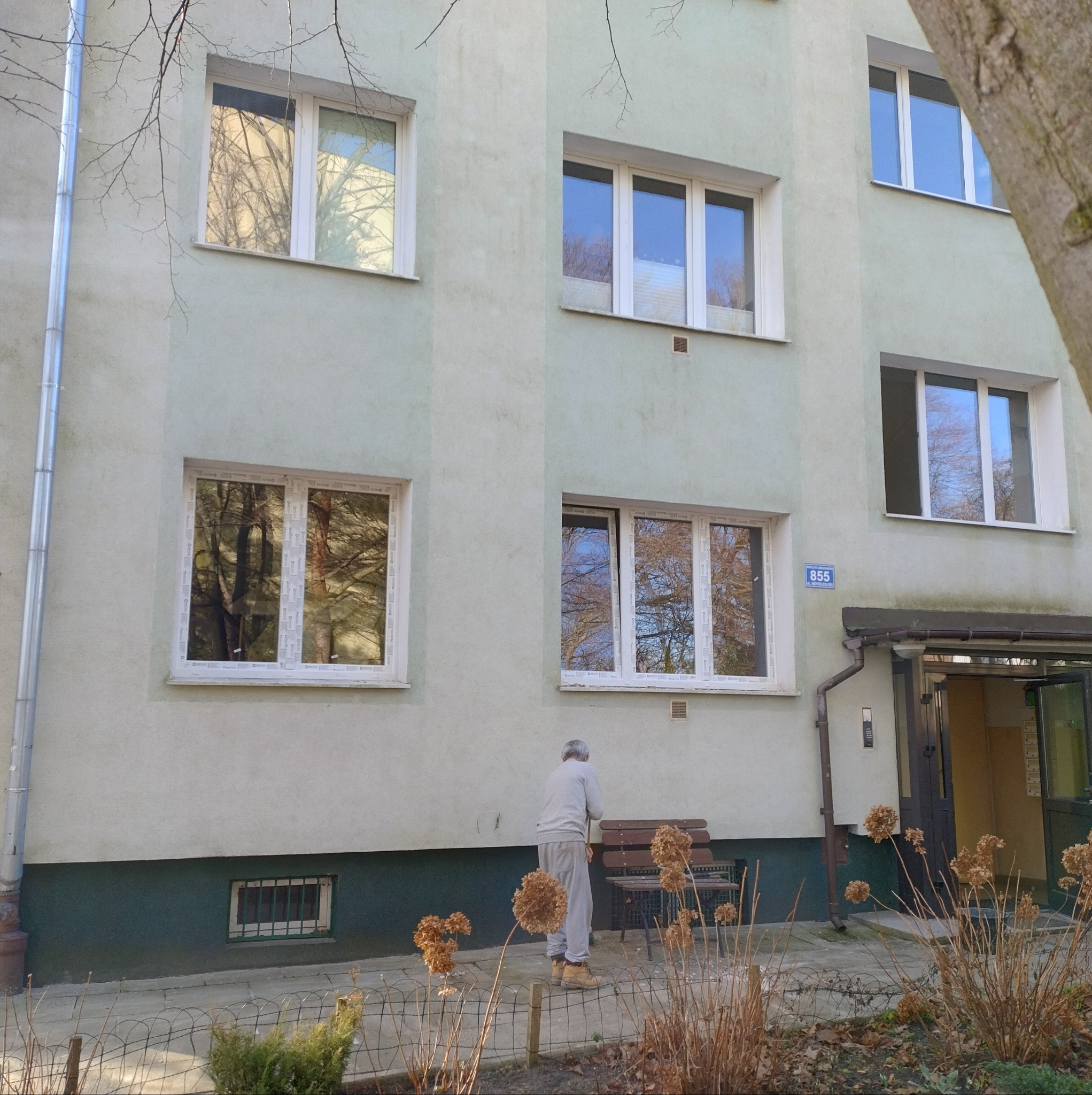 Budynek z oknami PCV, widok z zewnątrz. Na pierwszym planie starszy mężczyzna. W oknach odbija się krajobraz. Białe ramy okienne kontrastują z elewacją.