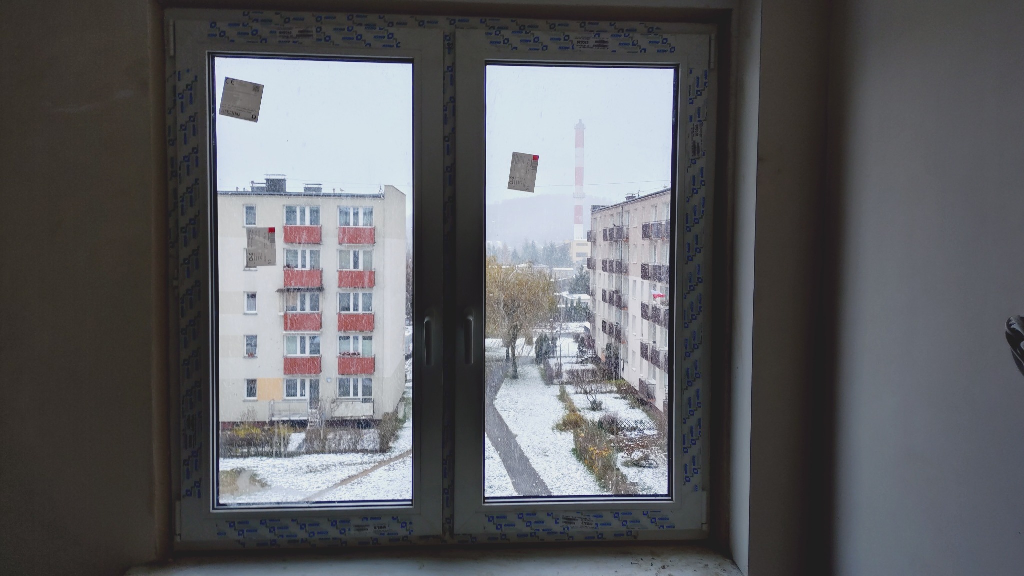 Nowe, dwuskrzydłowe okno PCV z widokiem na zimowy krajobraz miejski z blokami i kominem w tle. Na szybach widoczne naklejki producenta.