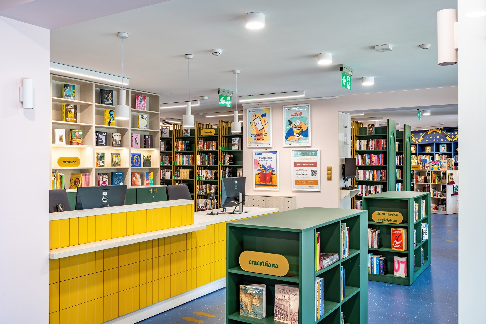 Żółto-zielona aranżacja wnętrza biblioteki z regałami pełnymi książek, stanowiskiem obsługi i plakatami informacyjnymi. Nowoczesny design z dbałością o detale.
