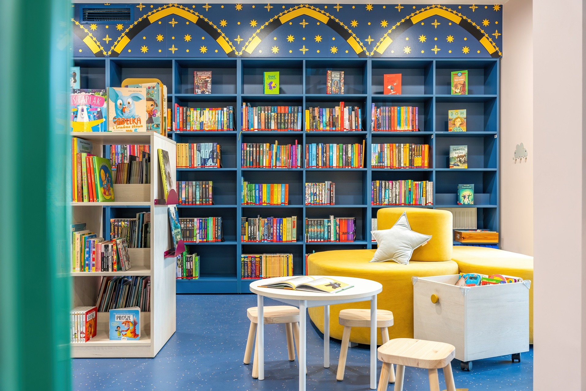 Aranżacja biura: kolorowa biblioteczka dla dzieci z książkami, stolikiem i pufami w odcieniach niebieskiego i żółtego, tworząca przyjazną przestrzeń do czytania.