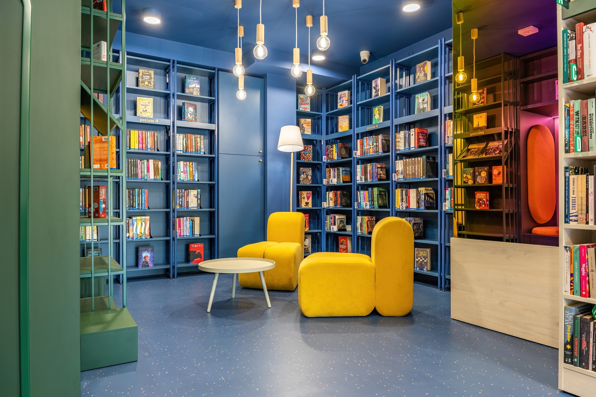 Niebieska biblioteka biurowa z żółtymi fotelami i stolikiem. Regały pełne książek, nowoczesne oświetlenie. Przytulna aranżacja przestrzeni biurowej.