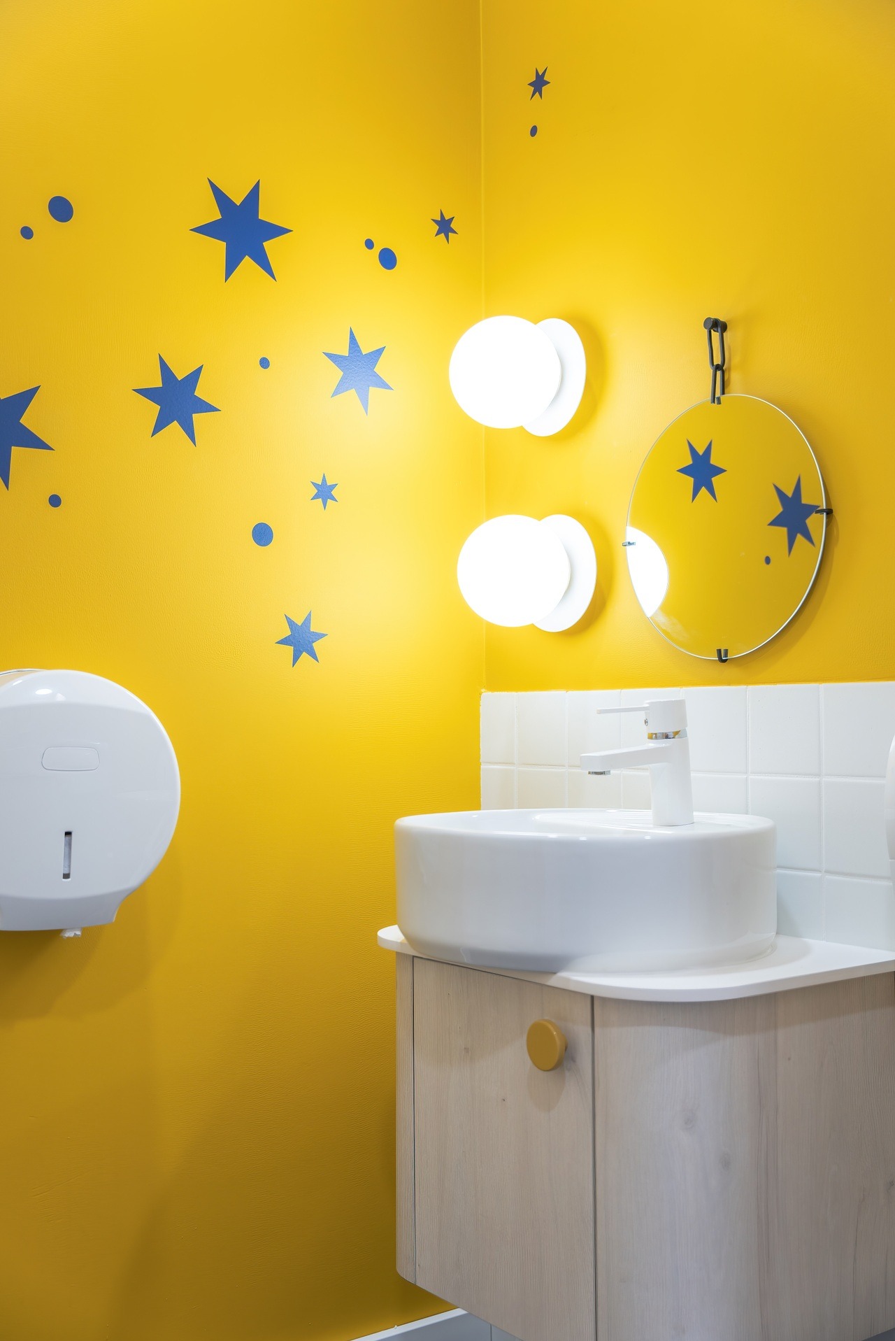 Żółta łazienka z niebieskimi gwiazdami: okrągłe lustro, biała umywalka na szafce z jasnego drewna, biały podajnik na ręczniki i okrągłe lampy.