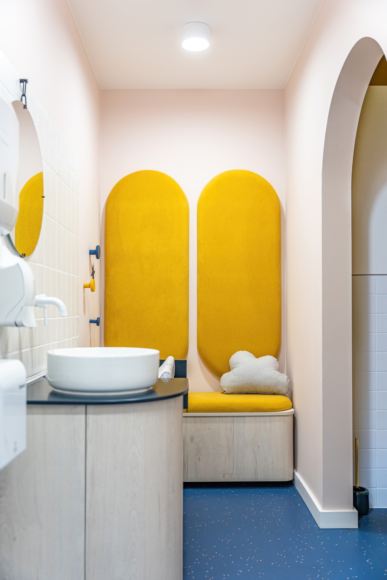 Nowoczesna łazienka biurowa z żółtymi panelami akustycznymi, umywalką na szafce z jasnego drewna i niebieską podłogą w kropki. Pastelowe ściany i designerskie dodatki.