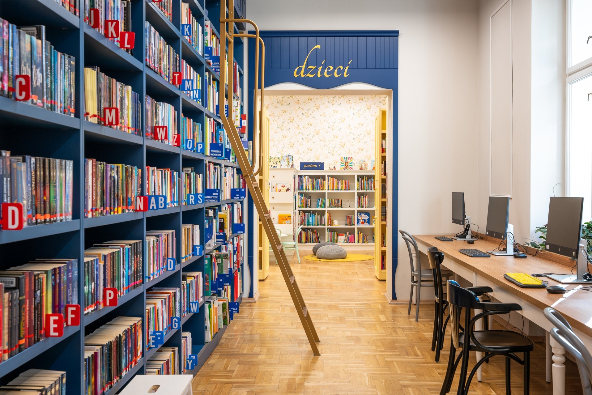 Jasne wnętrze biblioteki dla dzieci z regałami pełnymi książek, drabinką i stanowiskami komputerowymi. Parkiet na podłodze, niebieskie akcenty kolorystyczne.