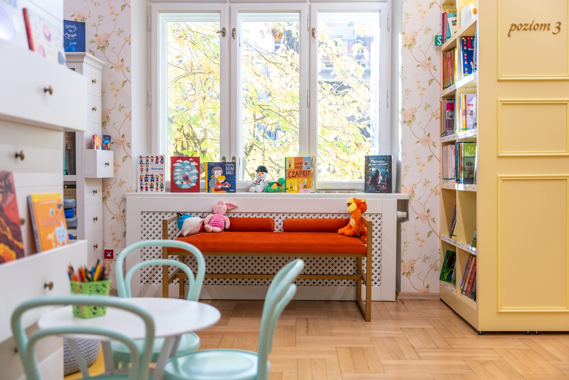 Jasne wnętrze biblioteki dziecięcej: regały z książkami, stolik z krzesłami, parapet z książkami i pluszakami. Tapeta w kwiaty i parkiet w jodełkę tworzą przytulny klimat.