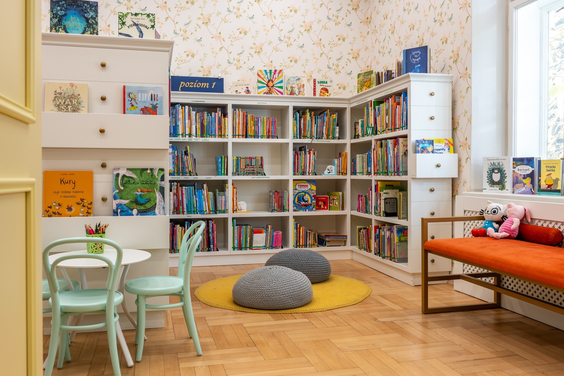 Jasne wnętrze biblioteki dla dzieci: białe regały z książkami, okrągły żółty dywan, miętowe krzesła i stolik. Tapeta w kwiaty i ptaki dodaje uroku.
