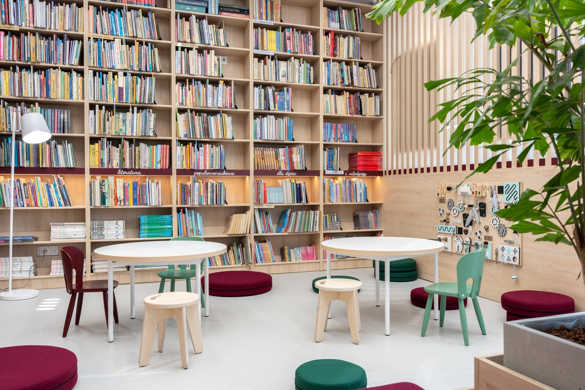 Jasne wnętrze biblioteki z regałami pełnymi książek, okrągłymi stolikami i krzesłami w pastelowych kolorach. Elementy dekoracyjne i rośliny dodają przytulności.