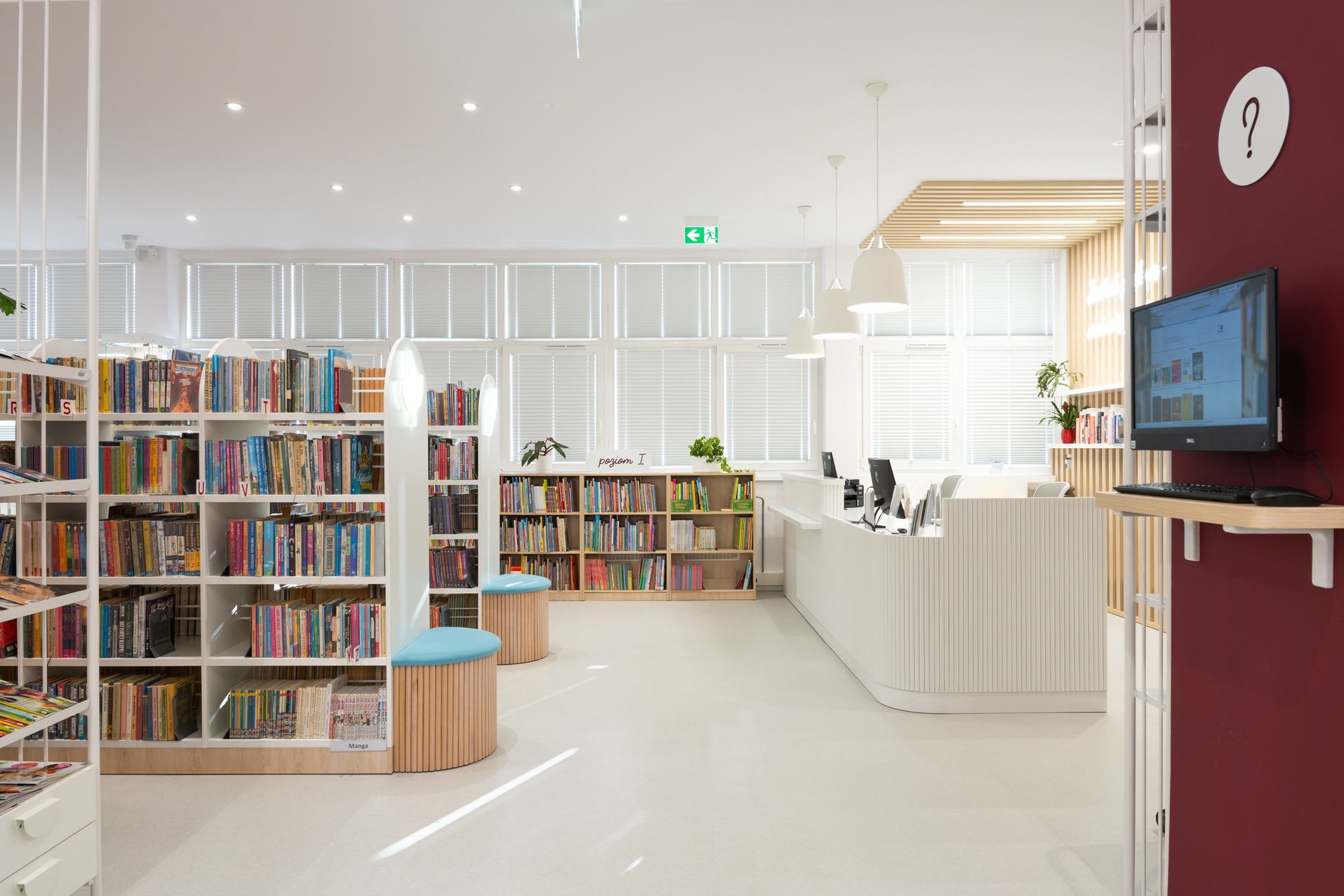 Jasne, nowoczesne wnętrze biblioteki z białymi regałami pełnymi książek, minimalistyczną ladą recepcyjną i elementami z naturalnego drewna. Widoczne stanowiska komputerowe.