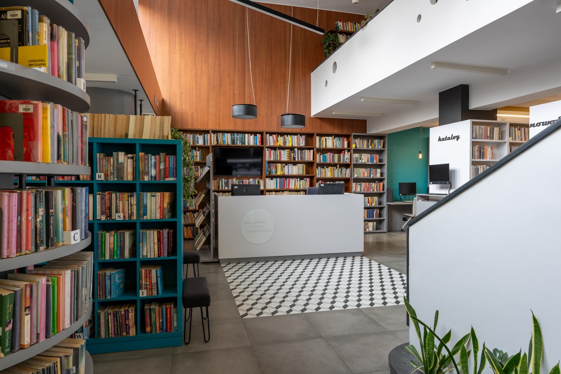 Nowoczesna biblioteka: regały z książkami, lada recepcyjna z napisem, wzorzysta podłoga, drewniane panele na ścianie i oświetlenie. Przestrzeń biurowa z książkami.