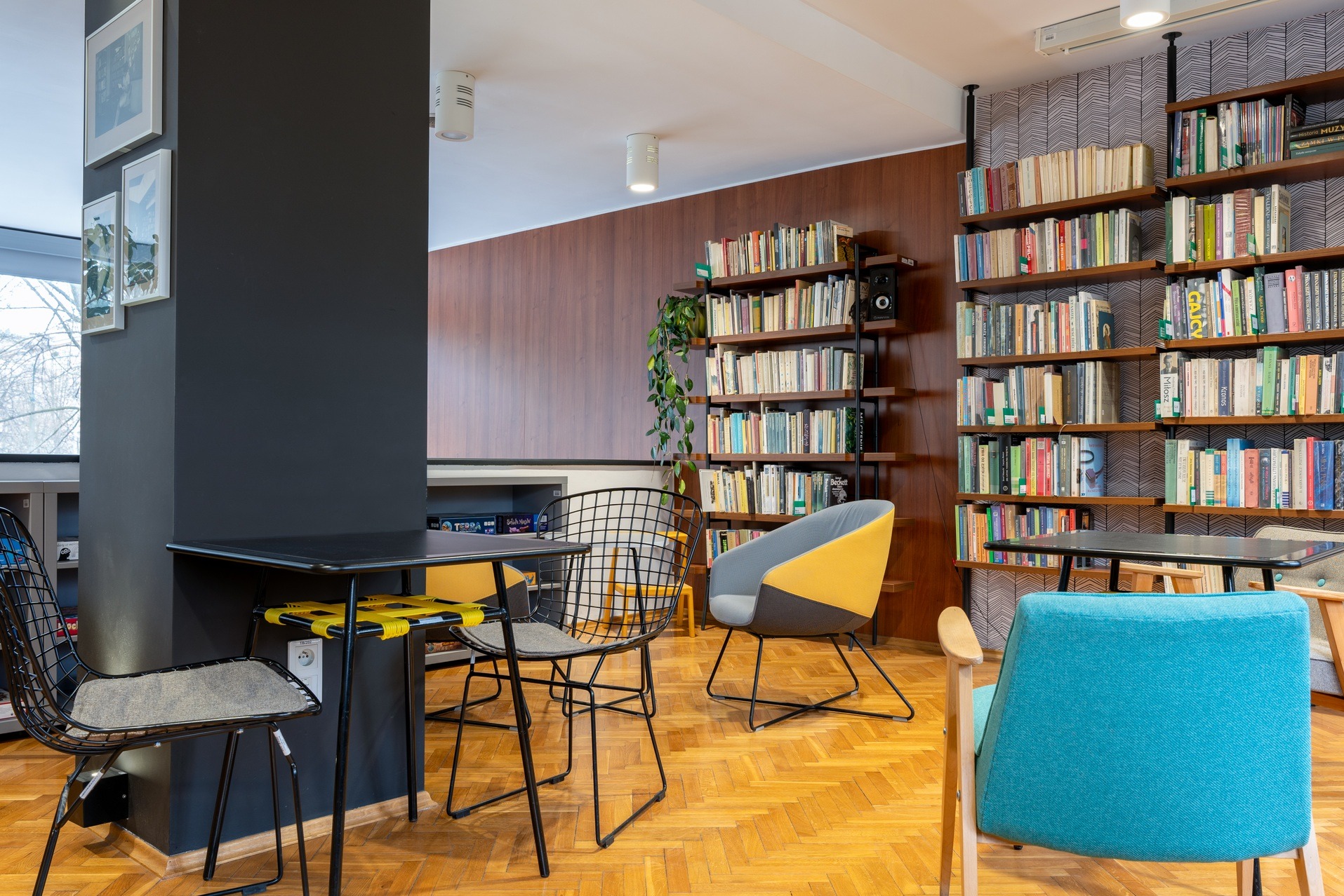 Stylowa aranżacja biura z regałami pełnymi książek, nowoczesnymi krzesłami i stolikami. Parkiet w jodełkę i designerskie oświetlenie tworzą przytulną atmosferę.