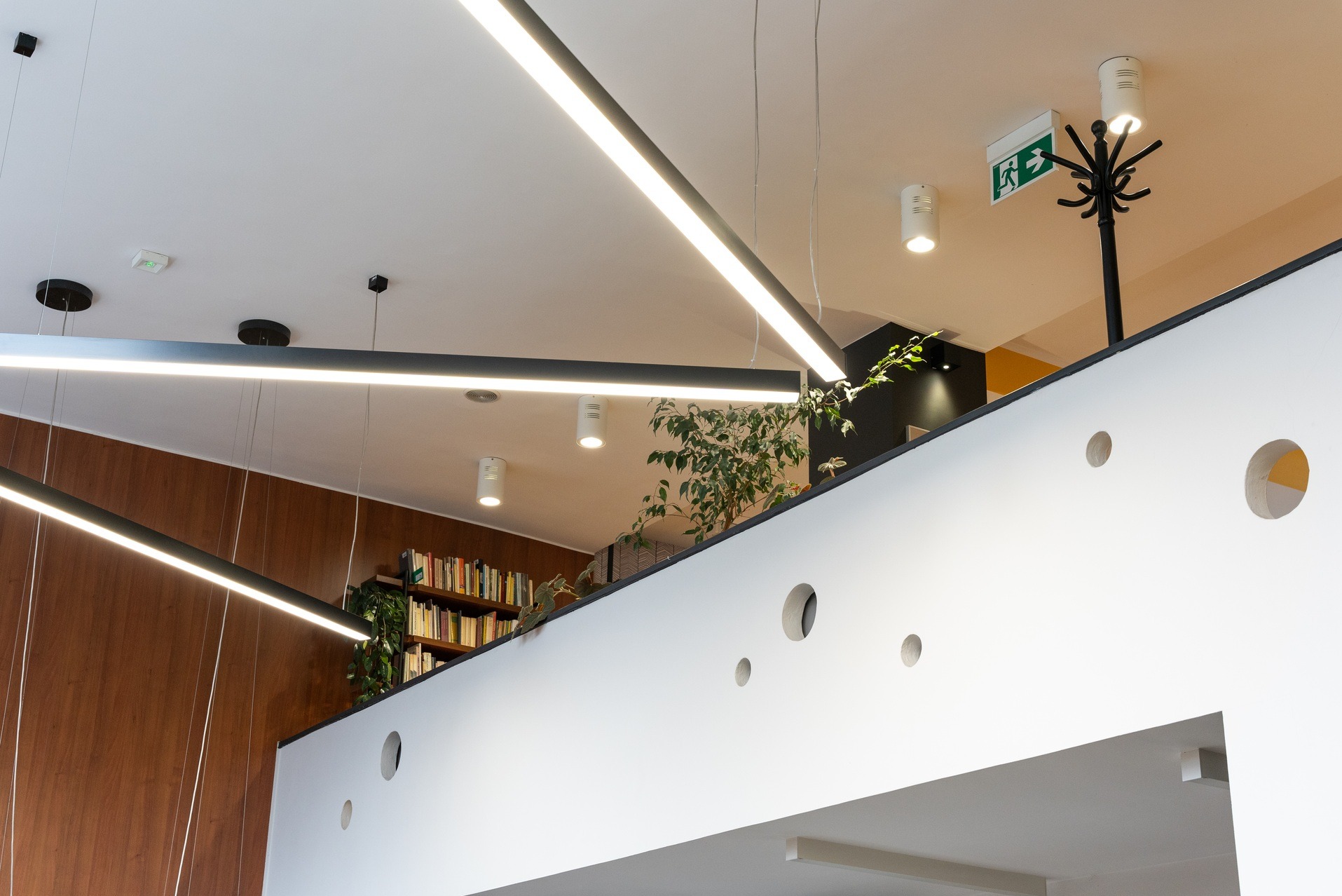 Nowoczesne biuro: biała ściana z okrągłymi otworami, oświetlenie liniowe LED, regał z książkami i rośliny. Widok z dołu, akcent na detale wykończenia.