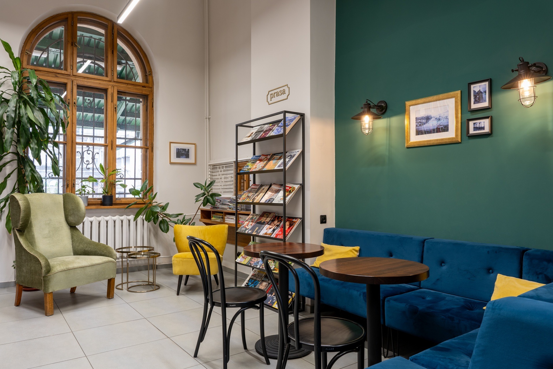 Stylowa aranżacja wnętrza biura z zieloną ścianą, niebieską sofą, drewnianymi stolikami i regałem z prasą. Widoczne rośliny doniczkowe i lampy w stylu retro.