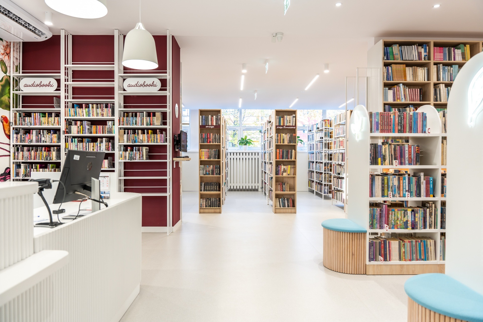 Jasne wnętrze biblioteki z regałami pełnymi książek, stanowiskiem obsługi klienta i elementami dekoracyjnymi w odcieniach bieli, bordo i błękitu. Nowoczesny design.