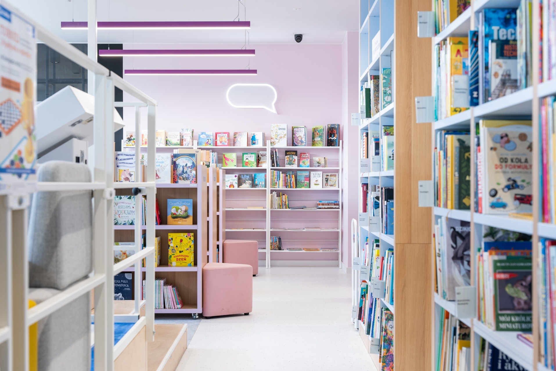 Jasne wnętrze biblioteki dziecięcej z różowymi akcentami: regały z książkami, pufy i neon w kształcie dymku. Nowoczesny design, pastelowa kolorystyka, przyjazna atmosfera.