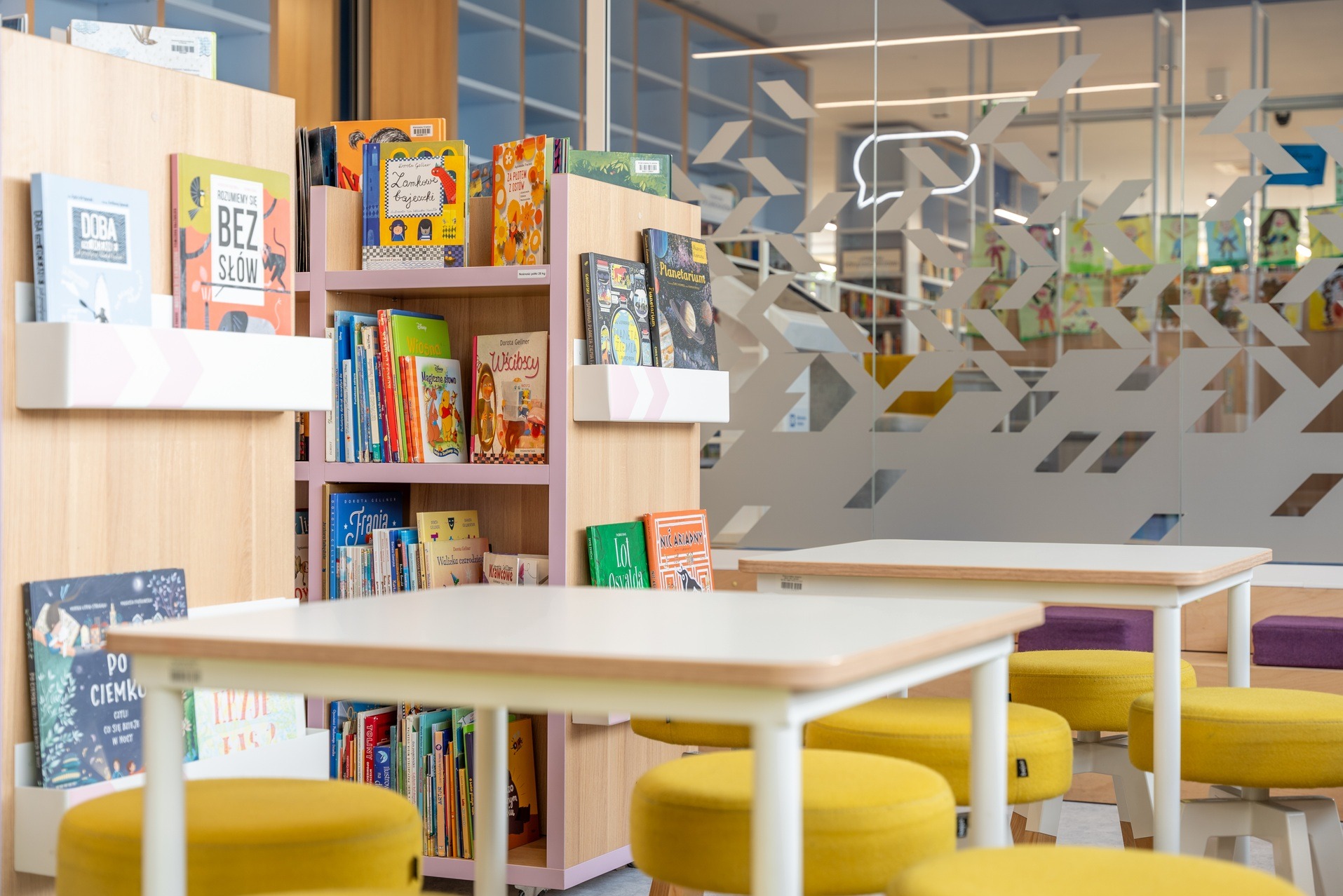 Jasne wnętrze biblioteki z regałami pełnymi książek dla dzieci, stolikami i żółtymi pufami. Na szklanej ścianie dekoracyjny wzór w kształcie strzałek i dymków.