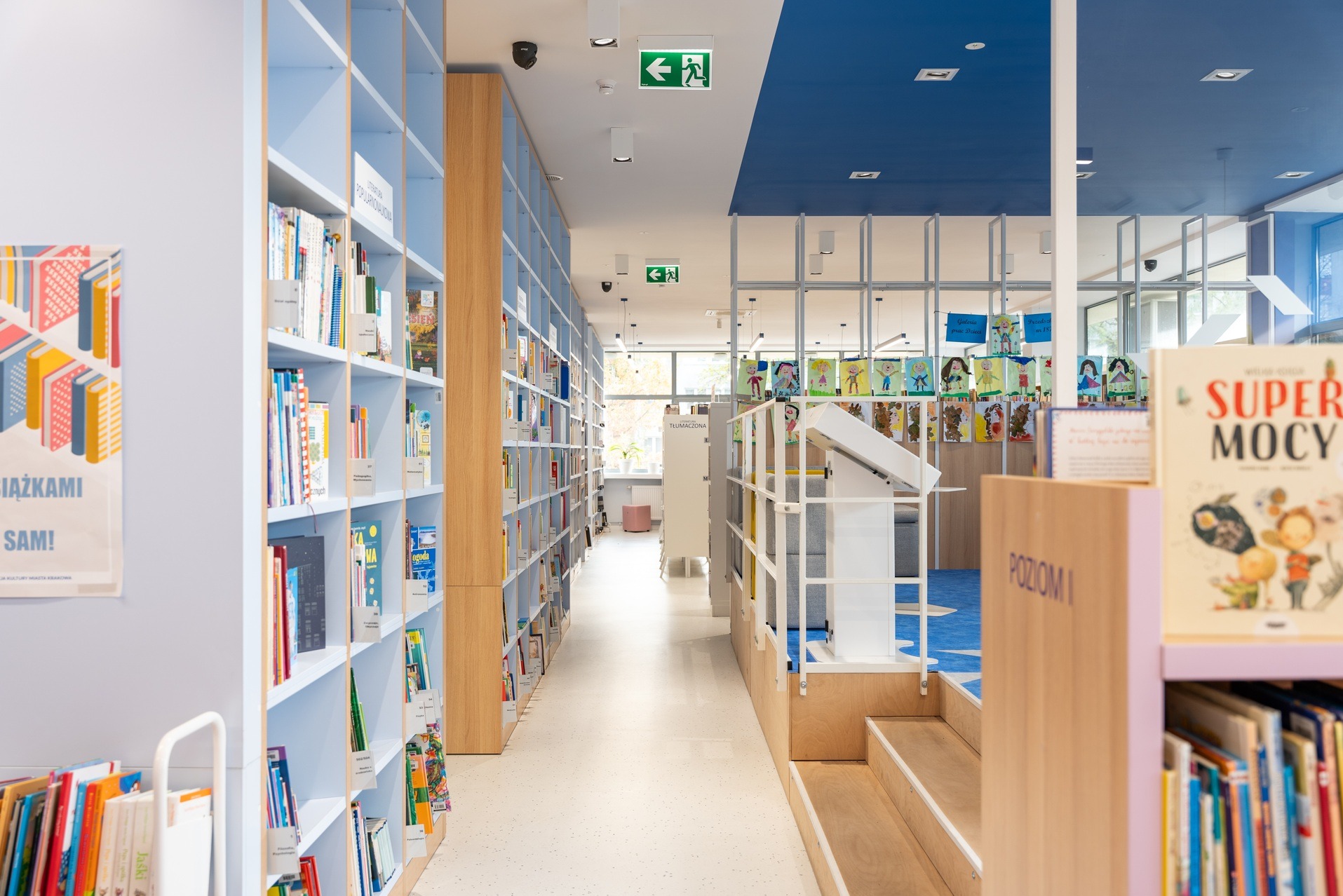 Jasne wnętrze biblioteki z regałami pełnymi książek, niebieskim sufitem i elementami wystroju dla dzieci. Widok w głąb pomieszczenia, z perspektywą na okno.