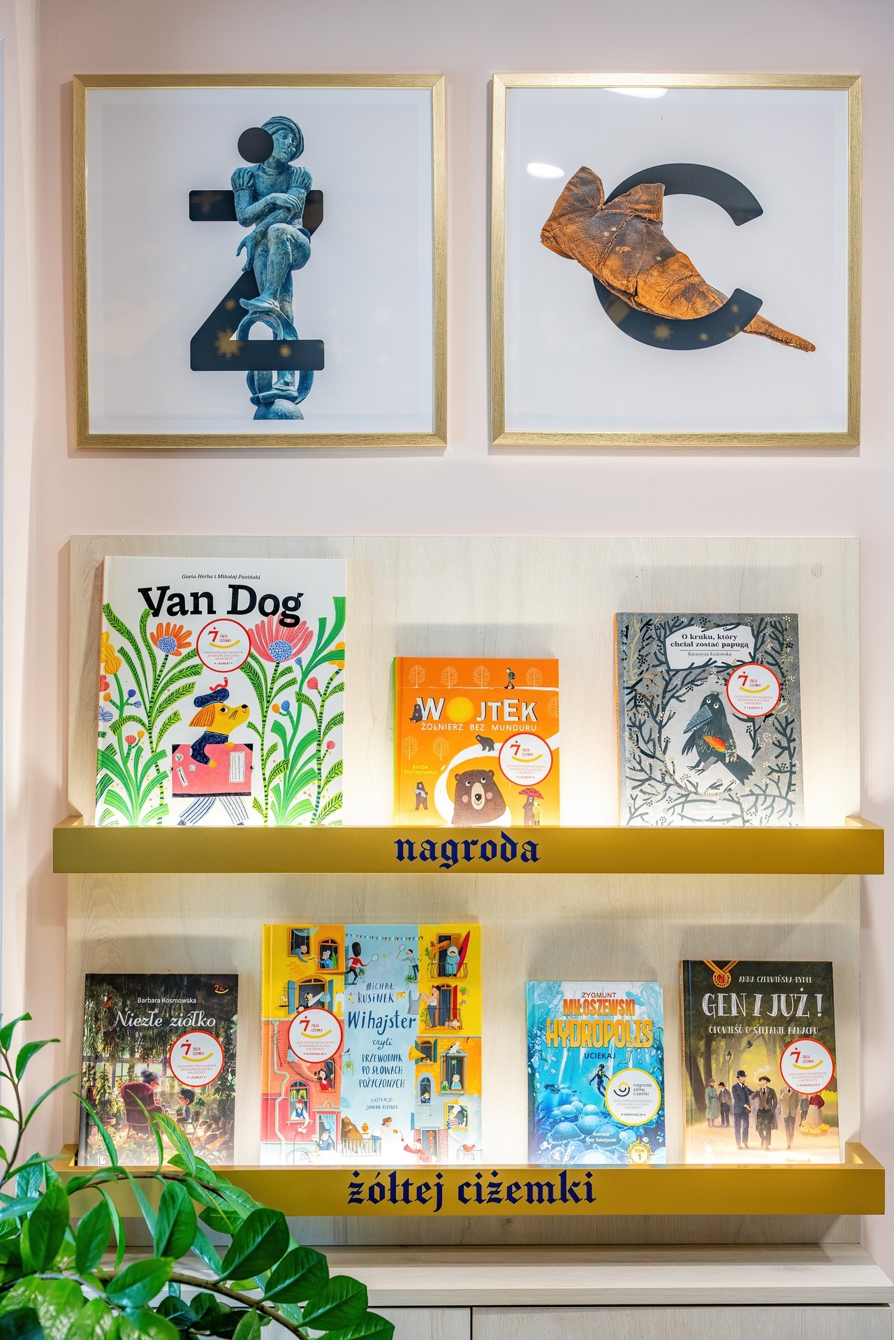 Kreatywna aranżacja ściany w biurze z półkami na książki dla dzieci, ozdobiona grafikami w złotych ramach i napisem 'nagroda żółtej ciżemki'.