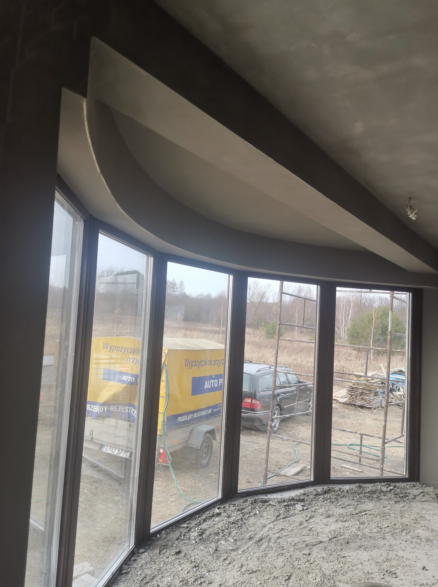 Wnętrze w trakcie prac: surowy tynk na ścianach i suficie z dekoracyjnym zaokrągleniem, duże okna z widokiem na zewnątrz. Widoczne rusztowania i materiały budowlane.